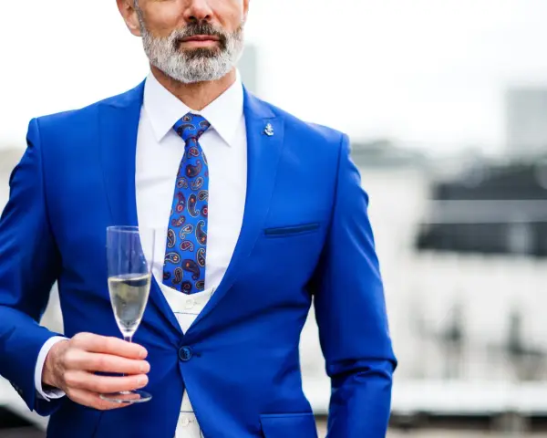 Elegancki mężczyzna w niebieskim garniturze i wzorzystym krawacie, trzymający kieliszek szampana. Idealny strój koktajlowy na specjalne okazje.