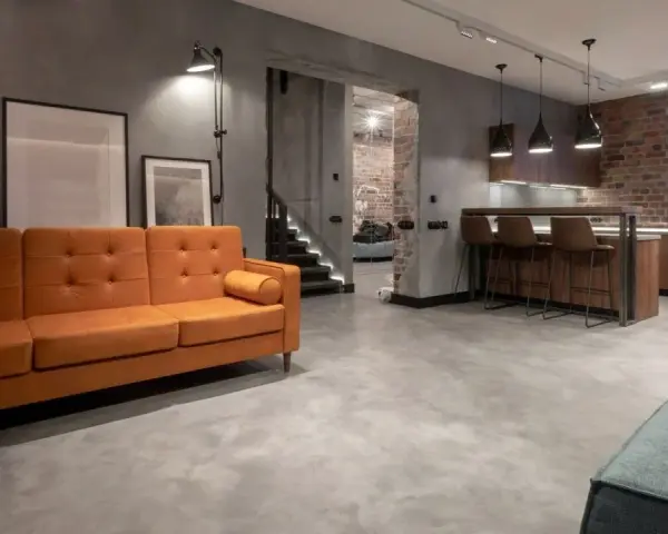 Nowoczesny salon z pomarańczową kanapą i kuchnią. Podłoga z mikrocementu na płytki nadaje industrialny charakter.