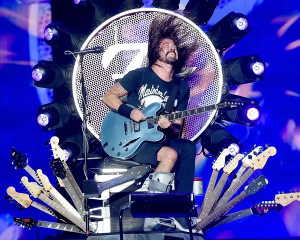 Dave Grohl z zespołem na scenie, szaleje z gitarą. Wokół niego mnóstwo gitar, a w tle wielkie logo i światła.