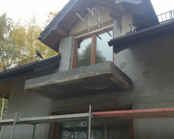 Budowa domu w jesiennej scenerii. Widać rusztowania, okna i balkon, a także ptaka na dachu. Trwają prace nad izolacją, która wpłynie na współczynnik przenikania ciepła.