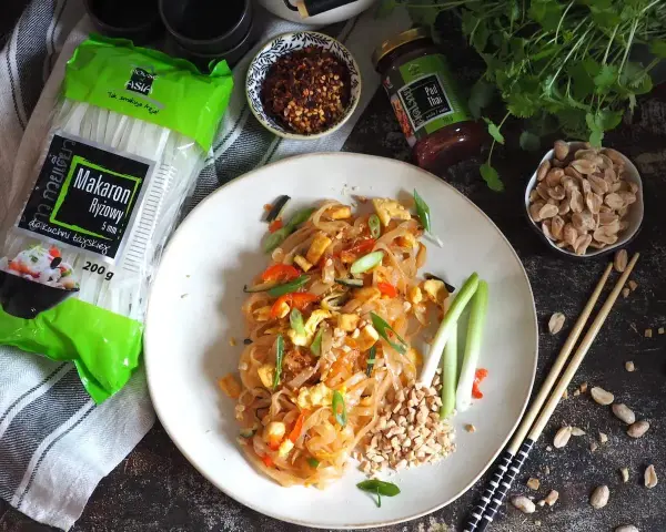 Gotowy Pad Thai przepis na talerzu, obok makaron ryżowy, orzeszki ziemne i sos.