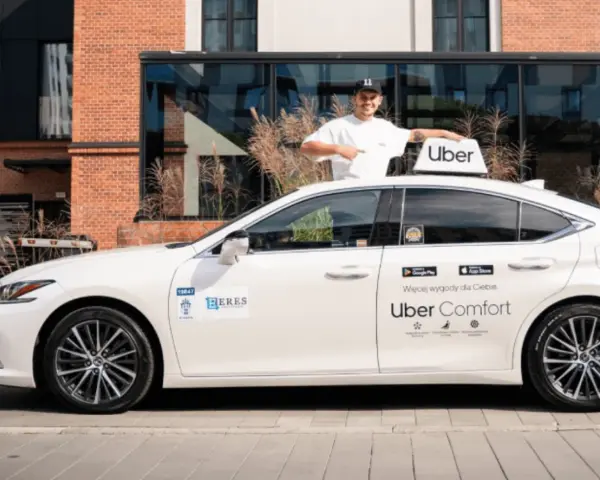 Mężczyzna w czapce z daszkiem stoi obok białego samochodu z logo Uber. To część floty Uber, gotowa do jazdy.