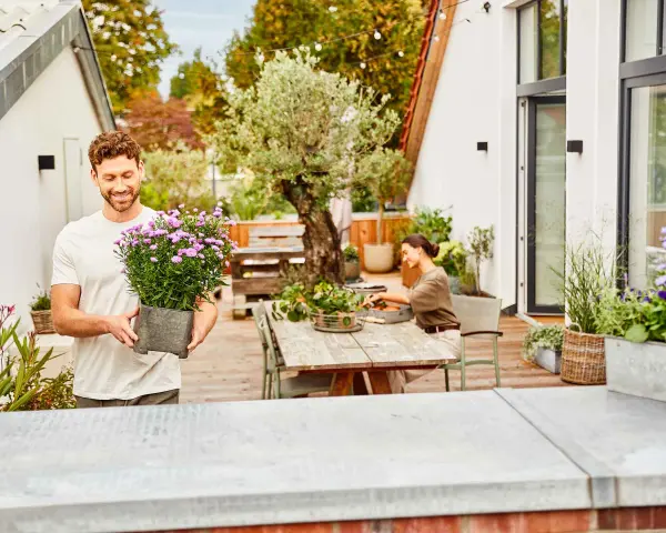 Mann hält Topf mit lila Blumen, um seine Dachterrasse mit Pflanzen zu gestalten. Eine Frau sitzt am Tisch.