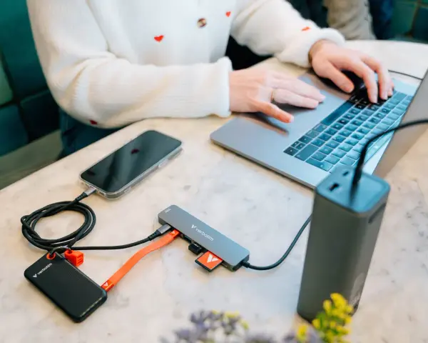 En person använder en laptop med en hubb ansluten till en telefon, extern hårddisk och powerbank. Hubben har usb 3.2-portar.