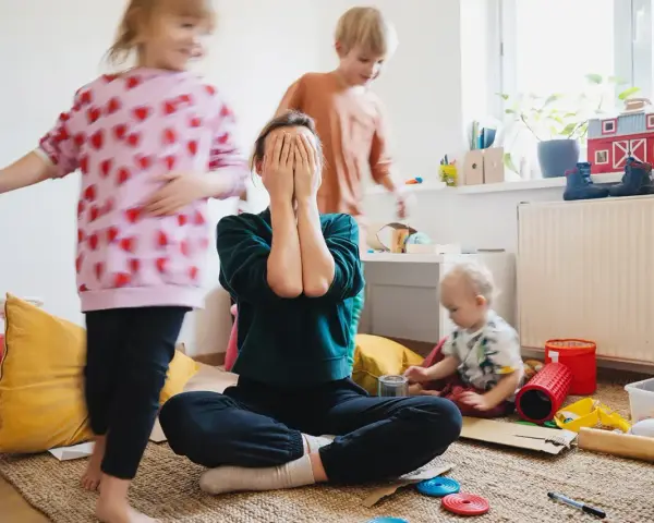 Chaos im Kinderzimmer. Eine Mutter versteckt ihr Gesicht in den Händen, während ihre Kinder spielen. Trotz des Trubels ist die liebe zu kindern spürbar.