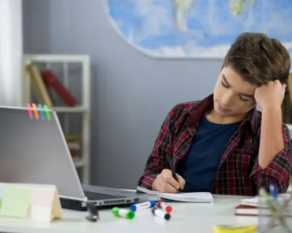 Ein Junge sitzt frustriert am Schreibtisch und macht seine hausaufgaben. Ein Laptop und bunte Stifte liegen bereit.