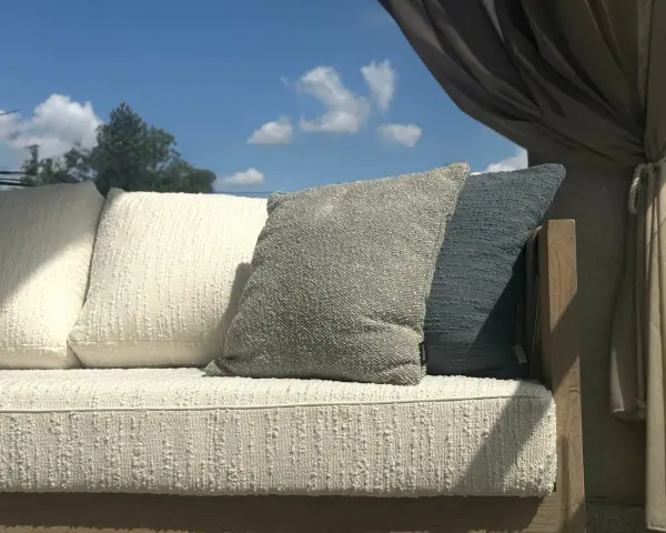 Wygodna sofa z poduszkami, osłonięta zasłonami, stoi na tle błękitnego nieba. Jej rama wykonana jest z pięknego, naturalnego **drewna tekowego**.