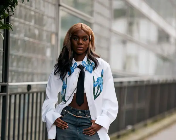 Modella con jeans baggy, un capo che simboleggia libertà e stile informale, indossa una camicia bianca con ricami floreali e cravatta.