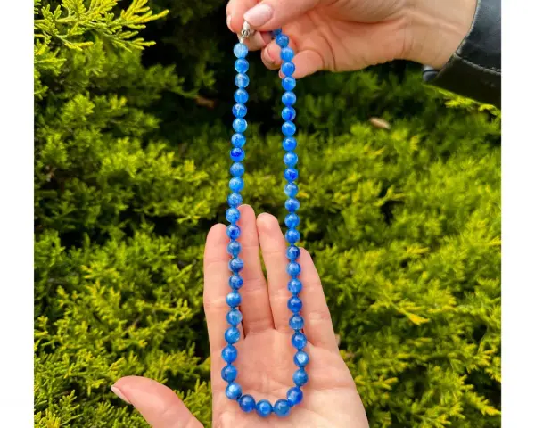 Collana di cianite blu, con perle luminose che catturano la luce, tenuta tra le mani su uno sfondo verde.