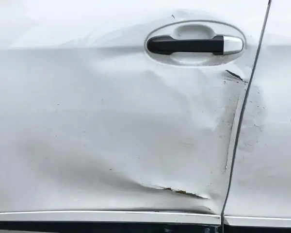 Widoczne wgniecenie i zarysowania na srebrnych drzwiach samochodu, świadczące o uderzeniu drzwiami na parkingu.