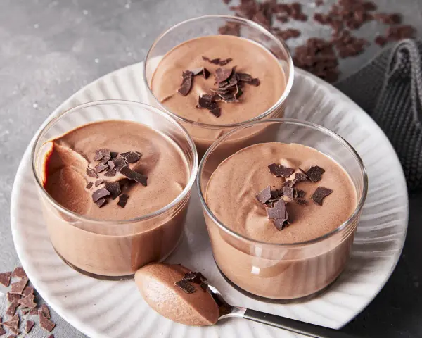 Drei Gläser mit luftiger Schokoladenmousse, garniert mit Schokostückchen. Ein Löffel zeigt die cremige Konsistenz dieses rezept mousse au chocolat ohne ei.