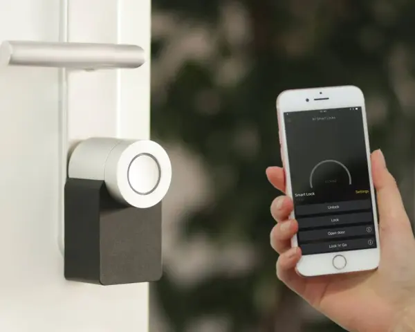 Mano sostiene un iPhone que muestra una app para controlar un cerrojo inteligente. La puerta tiene un diseño moderno.