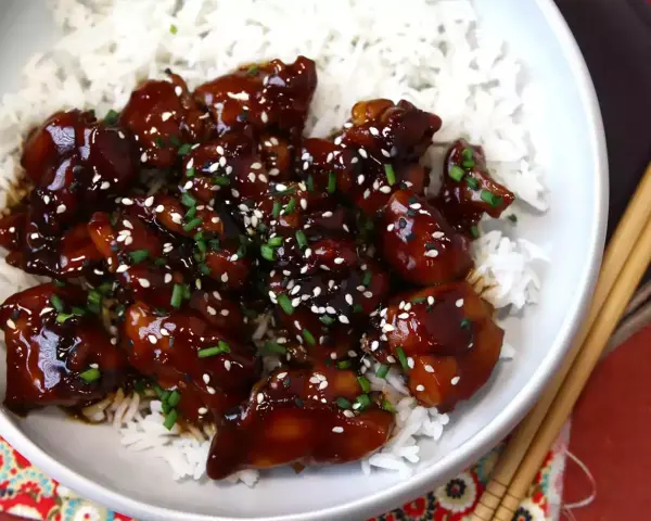 Trozos de pollo teriyaki glaseados y brillantes, cubiertos con semillas de sésamo y cebollino, servidos sobre una cama de arroz blanco.