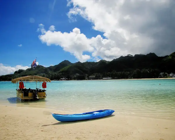 A blue kayak rests on a sandy beach in the Cook Islands, with a thatched-roof boat and lush green mountains in the background.