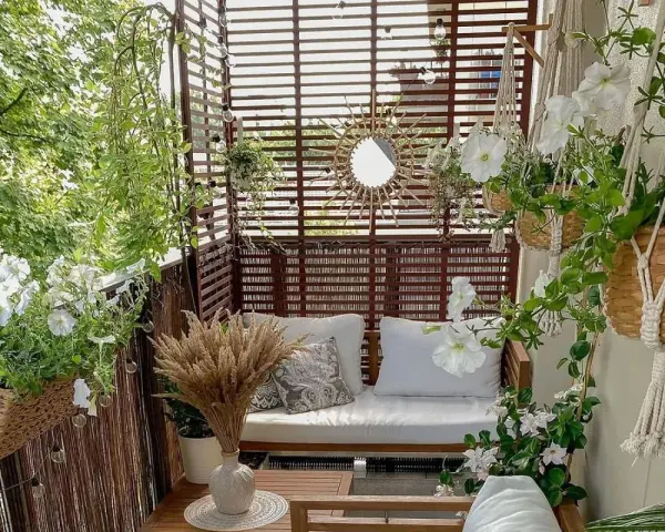 Przytulny balkon z drewnianą sofą, białymi kwiatami i ozdobnym lustrem w kształcie słońca.