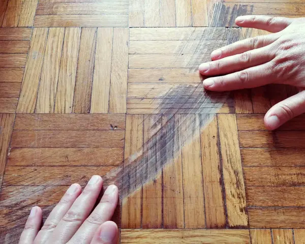 Mani sul parquet rovinato, mostrando i graffi e le macchie. Si valuta come pulire il parquet rovinato per riportarlo al suo antico splendore.
