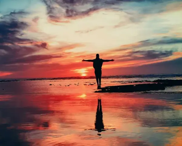 Człowiek z rozłożonymi ramionami na plaży o zachodzie słońca. To jakby szukał odpowiedzi na pytanie: asceza co to?