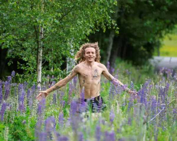 En man med lockigt hår och tatueringar står i en äng full av lila lupiner. Han ser ut att njuta av naturen, kanske som en del av ett avsnitt av "spring snyggt podden".