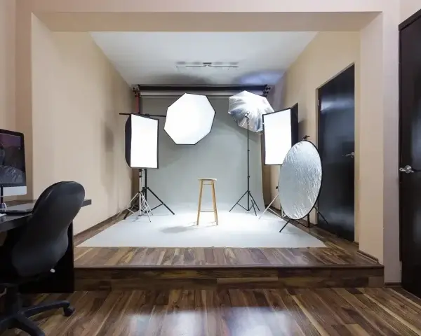Domowe studio fotograficzne z oświetleniem, statywami, blendą i krzesłem. Po lewej komputer z monitorem.
