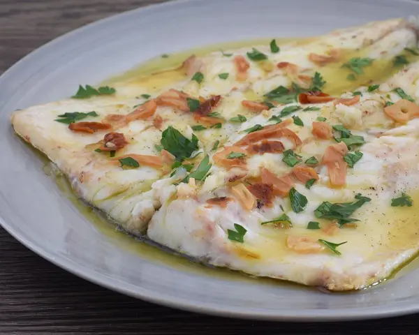 Pescado blanco cocinado en aceite con ajo laminado y perejil picado, servido en plato gris.