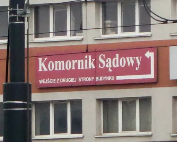 Tablica "Komornik Sądowy" na budynku. Wskazuje wejście z drugiej strony.