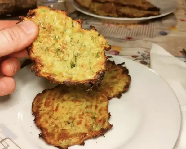 Una mano tiene una frittella di zucchine dorata e croccante. Sul piatto, altre frittelle di zucchine invitano ad essere gustate.