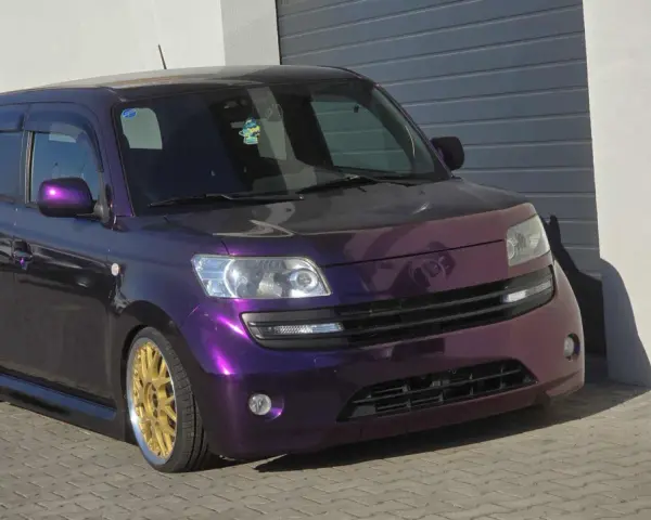 Fioletowy Daihatsu Materia z błyszczącymi złotymi felgami, zaparkowany przed garażem.