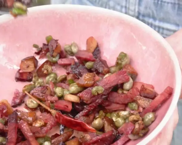 Un plato rosa lleno de habas con jamón, guisantes y un chile rojo seco.