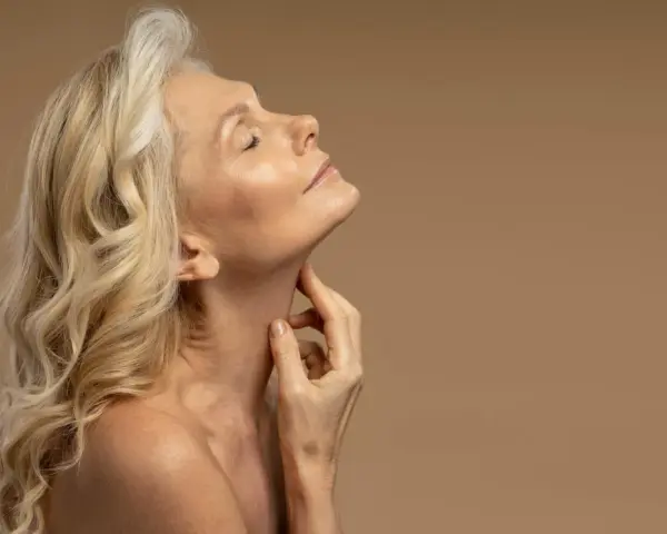 Kobieta z blond włosami i zamkniętymi oczami delikatnie dotyka szyi. Jej skóra w okresie menopauzy jest widoczna, podkreślając naturalne piękno.