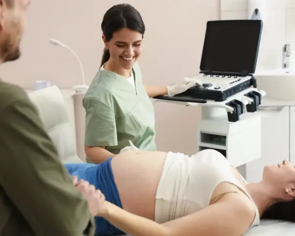 Uśmiechnięta lekarka wykonuje badanie wolnego płodowego dna u ciężarnej pacjentki, podczas gdy partner wspiera ją, trzymając za rękę.