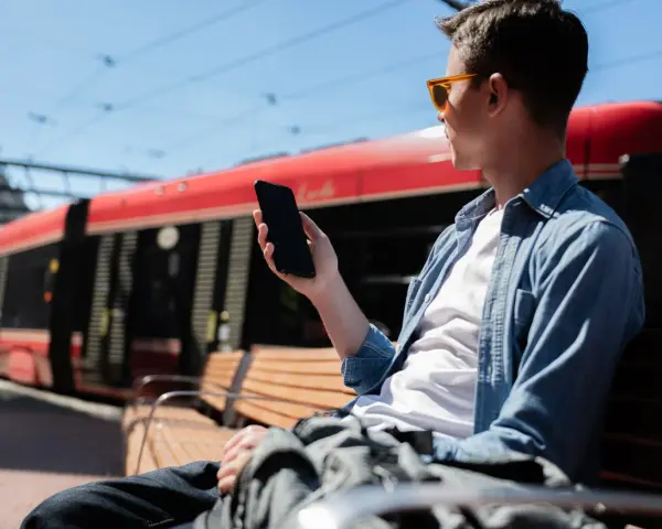 Mężczyzna w okularach siedzi na ławce i trzyma telefon. W tle czerwony tramwaj.