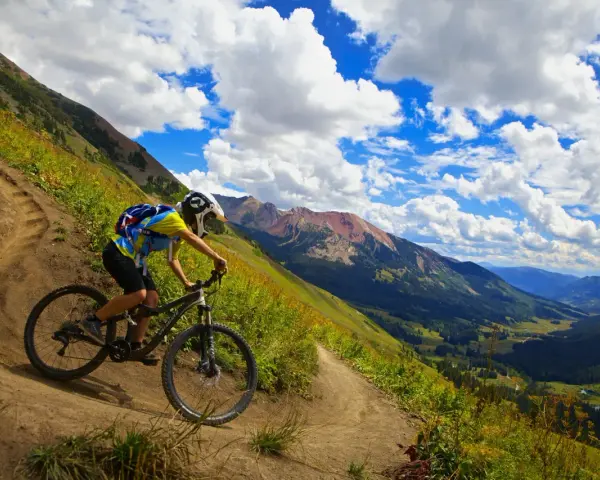 A mountain biker navigates a winding trail, enjoying the breathtaking views of cross country mountain bike trails winding through a lush valley and rugged peaks under a dramatic sky.