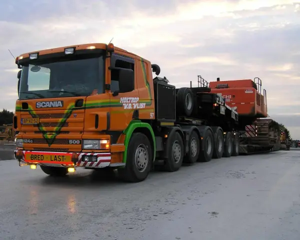 Pomarańczowy Scania z naczepą przewozi koparkę. Transport ponadgabarytowy wymaga specjalnych godzin jazdy.
