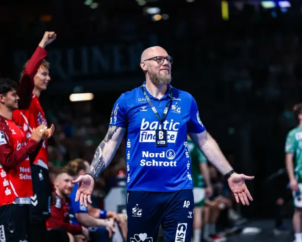 Trener SG Flensburg-Handewitt w niebieskiej koszulce z tatuażem na ramieniu, z otwartymi dłońmi, podczas meczu.