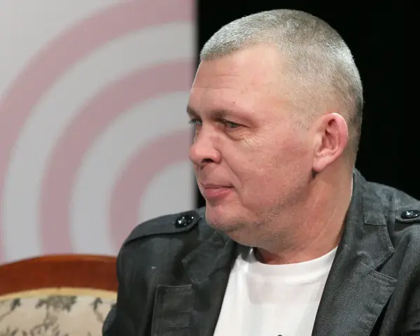 Adam z zespołu Raz Dwa Trzy w szarej marynarce, siedzi na kanapie.