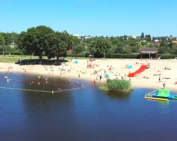 Słoneczny dzień nad jeziorem Grzymisławskim. Ludzie odpoczywają na plaży, kąpią się i bawią na dmuchanych atrakcjach.