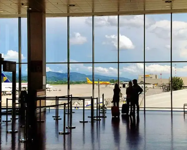Widok z okna na lotnisko Bergamo. Trzy osoby z walizkami czekają na samolot. W tle żółty samolot DHL i góry.