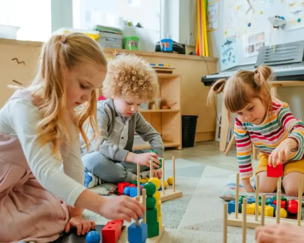 Drei Kinder, darunter ein **kind 8 jahre**, spielen konzentriert mit bunten Holzbausteinen im Kindergarten.