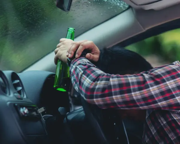 Mężczyzna w kratę, z butelką w ręku, opiera się o kierownicę. Pytanie: po ilu latach występuje przedawnienie za jazdę pod wpływem alkoholu?