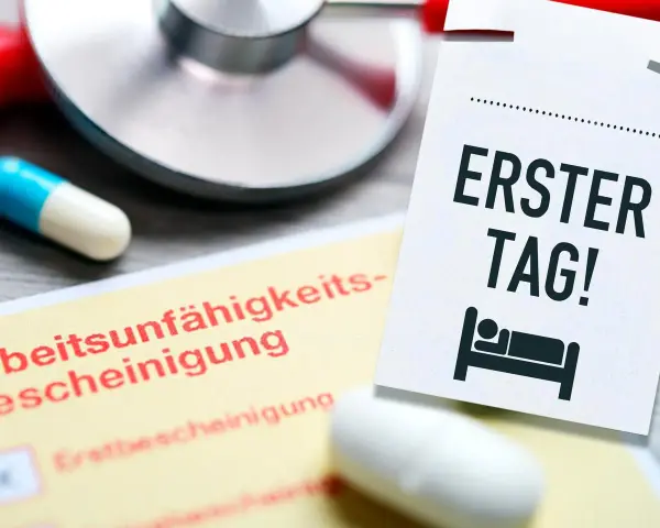 Arbeitsunfähigkeitsbescheinigung und eine Notiz "Erster Tag!" mit einem Bett-Symbol. Ein Stethoskop und Pillen deuten auf eine Krankschreibung hin, um gesund schreiben zu lassen.