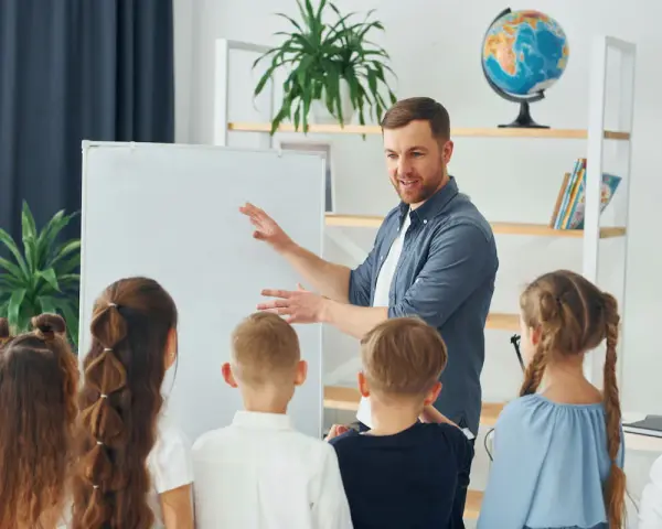 Nauczyciel tłumaczy coś dzieciom przy tablicy. To mogą być dni dyrektorskie, podczas których uczniowie poznają szkołę.