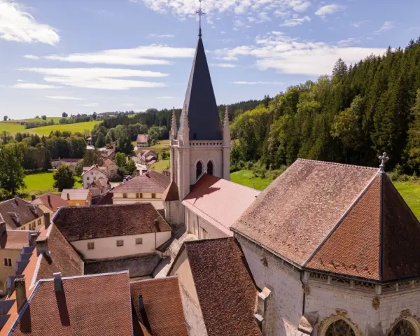 Vue aérienne d'un village pittoresque du haut Doubs, dominé par une église à la flèche élancée, entouré de collines verdoyantes et de forêts.