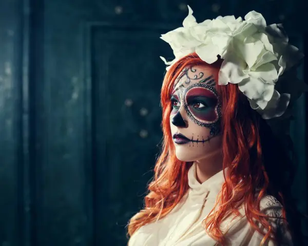 Kvinna med detaljerad ansiktsmålning för halloween, klädd i en vit blus och med en stor blomsterkrans.
