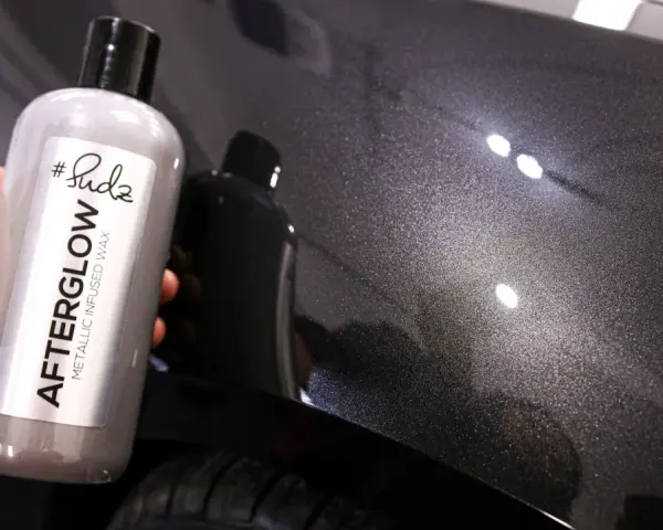 Bottle of "Afterglow Metallic Infused Wax" held against a dark, metallic car finish, showcasing the product's ability to create multiple coats of wax for a deep shine.