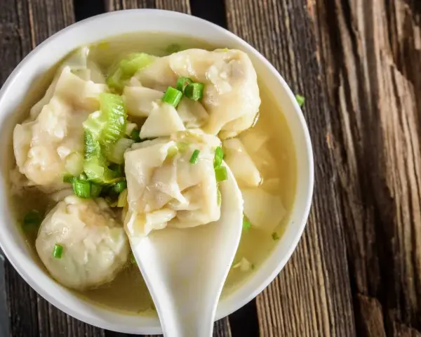 Aromatyczne pierożki wonton w bulionie, ozdobione szczypiorkiem i kawałkami warzyw, na drewnianym tle.