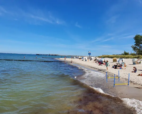 En solig tysk strand med folk som njuter av havet och sanden. En brygga sträcker sig ut i vattnet.