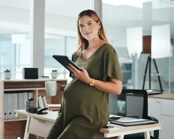 Uśmiechnięta ciężarna kobieta z tabletem i kubkiem w biurze. Dowiedziałam się o ciąży po okresie wypowiedzenia, ale teraz planuję przyszłość.