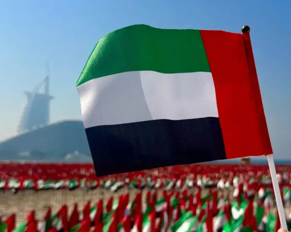 Flaga Zjednoczonych Emiratów Arabskich powiewa na tle drapaczy chmur w Dubaju. W tle widać morze małych flag.