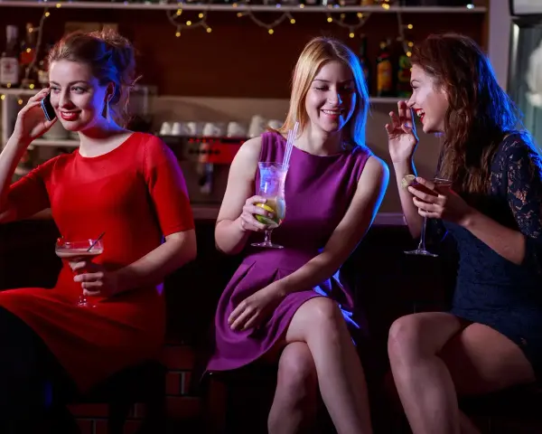 Trzy kobiety w **koktajlowych kreacjach** siedzą przy barze, pijąc drinki i rozmawiając.