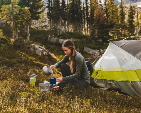 A woman enjoys a peaceful moment of camping alone, preparing a warm drink next to her tent amidst a serene forest landscape.